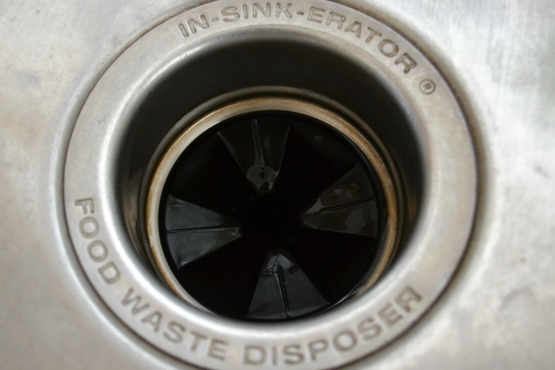 Top-down view into a stainless kitchen sink's InSinkErator disposal opening, showing the black rubber splash guard and staining around the rim