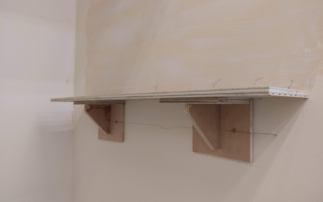 Wooden shelf mounted on two wooden wall brackets with a pencil layout line visible on the drywall behind it