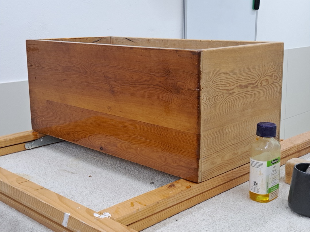 A wooden storage box partway through refinishing, with a freshly oiled section showing deep color next to bare sanded wood, and a bottle of finishing oil beside it