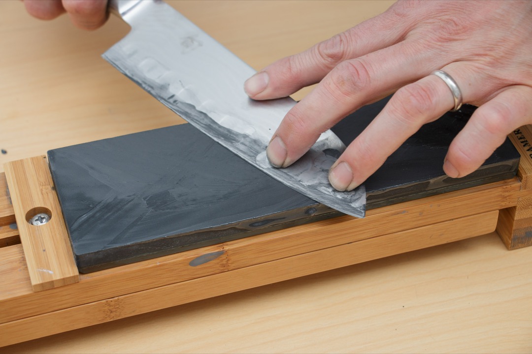A chef's knife being drawn across a wet whetstone at a low angle, water pooling on the stone's surface, demonstrating the whetstone sharpening technique covered in this guide