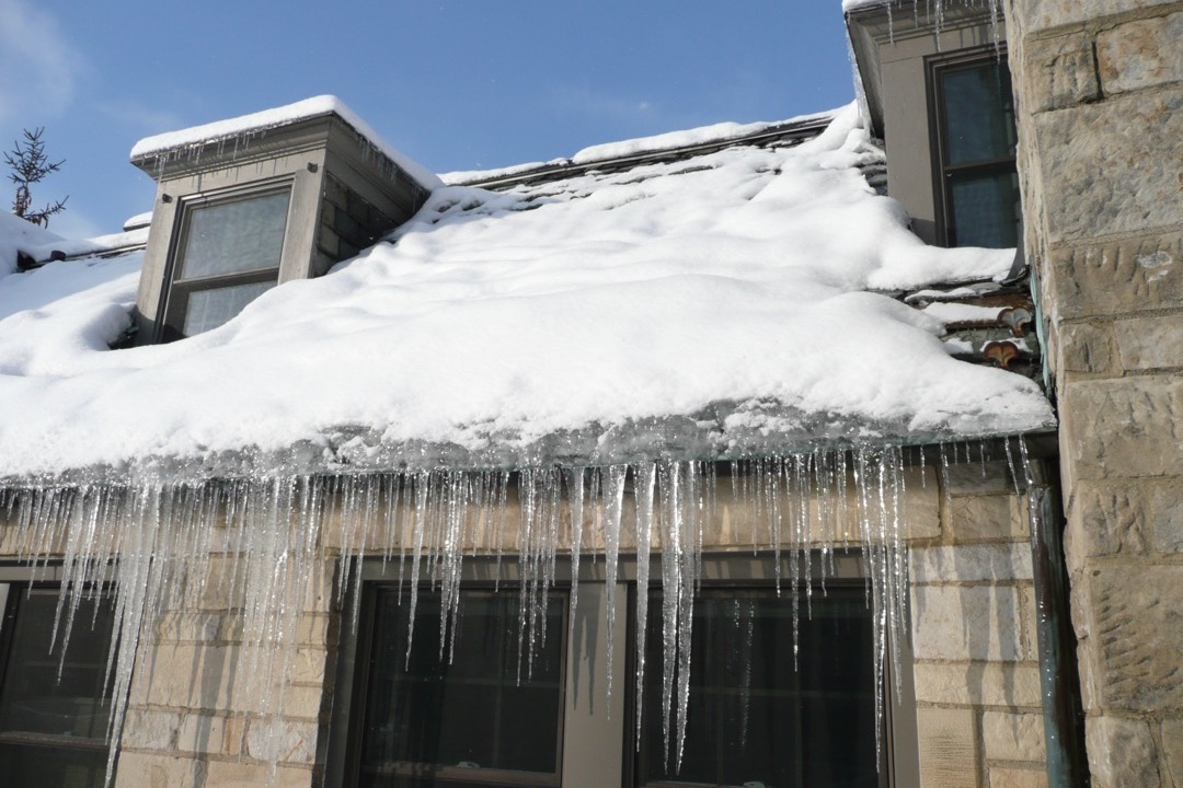 Snow-covered residential roof with icicles forming at the eaves and gutters showing visible ice dam buildup, illustrating the winterization risks this guide prevents