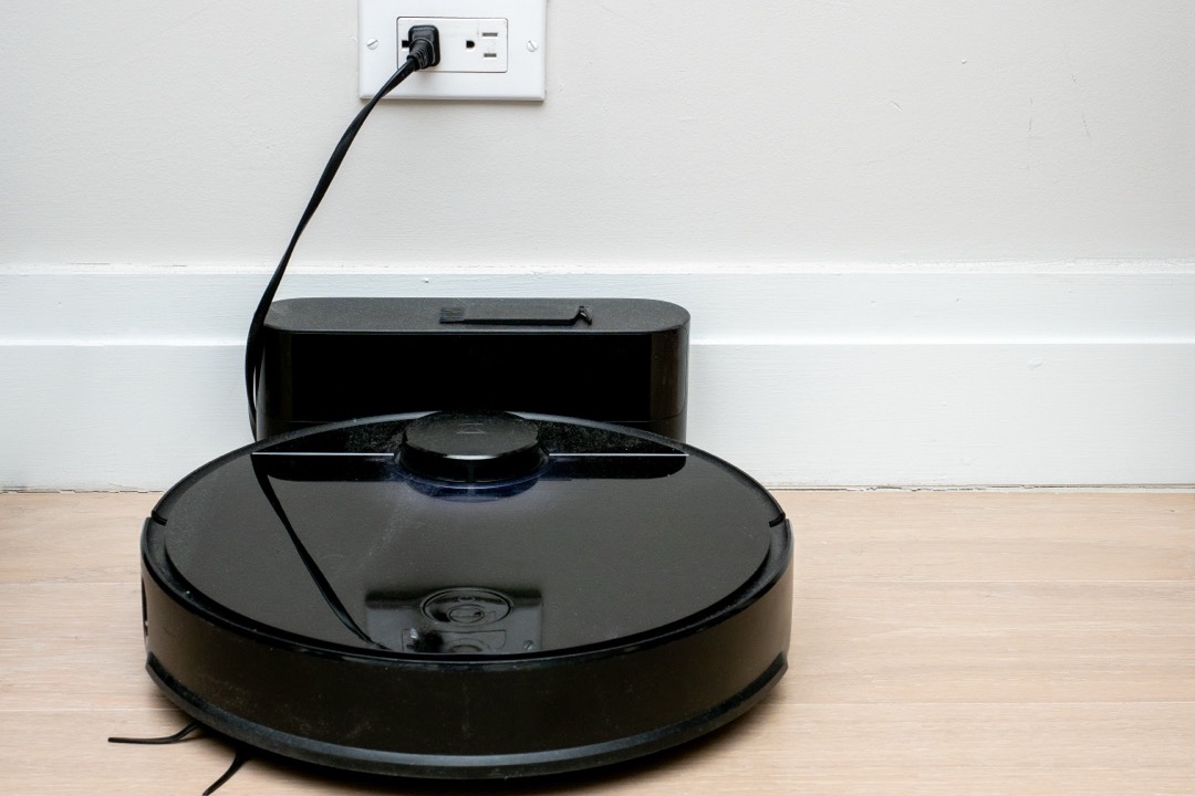 A robot vacuum cleaning a hardwood floor next to a stick vacuum standing upright against a wall